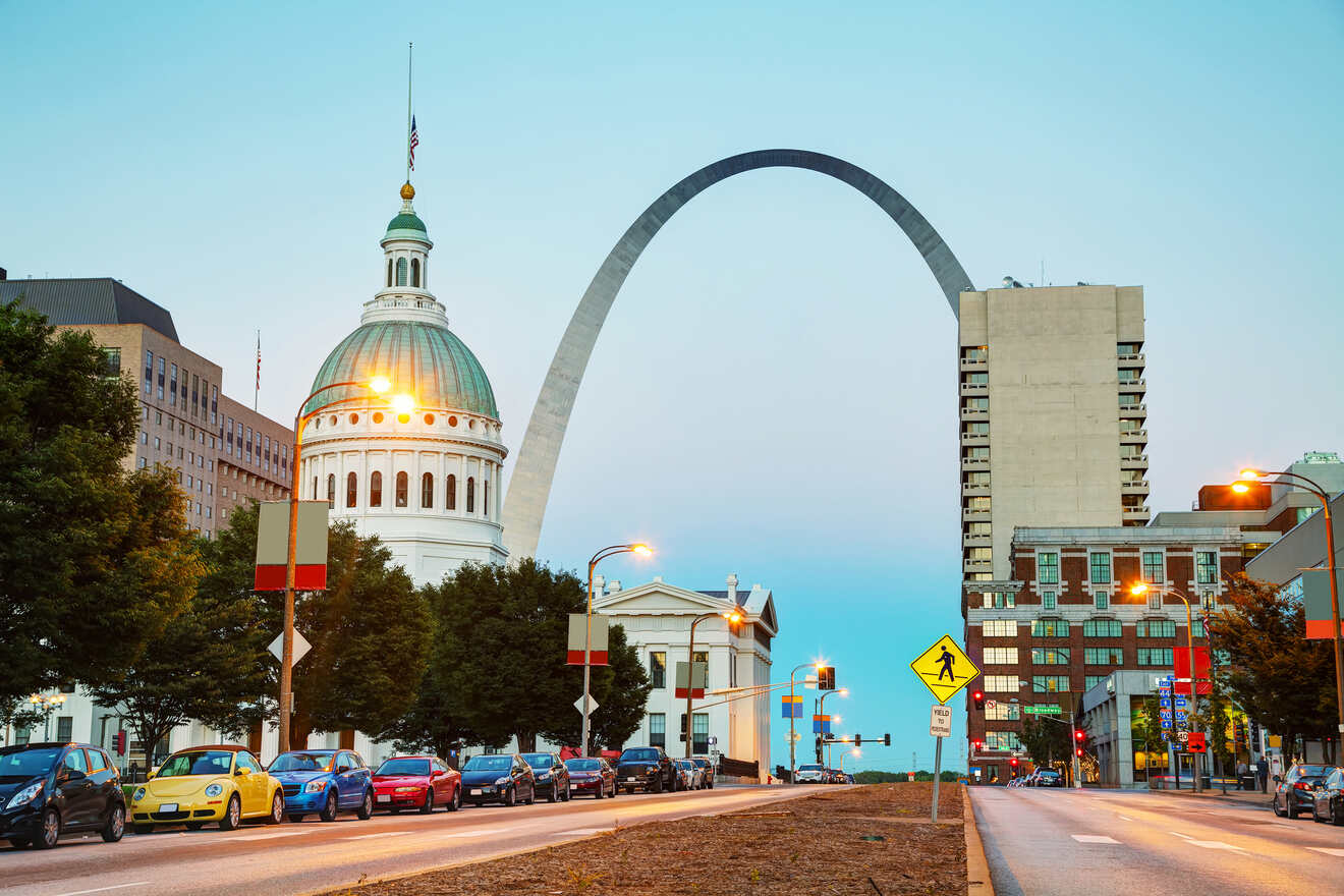 Twilight view of a St Lousi Missouri cityscape, with a distinctive arch monument and domed building, as seen from a street lined with cars